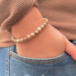 May include: A beaded bracelet with light brown and beige spherical beads. The bracelet is worn on a wrist, with a brown sweater sleeve and blue jeans visible in the background.