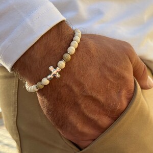May include: A white and brown beaded bracelet with a silver cross charm. The bracelet is worn on a person's wrist.
