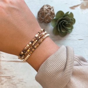 May include: A set of three bracelets with brown, white, and gold beads. The bracelets are stacked on a person's wrist.