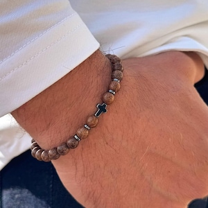 Puede incluir: Un brazalete de cuentas de madera marrón con un dije de cruz de metal negro.