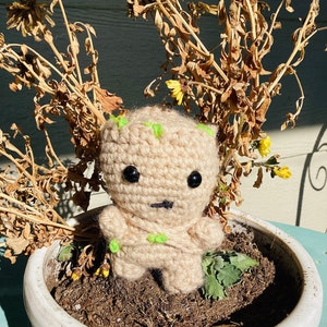 May include: A crocheted Groot character in a beige color with green accents. The character is sitting in a white ceramic pot with brown soil and dried flowers.