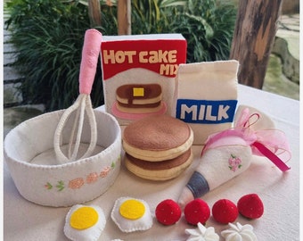 Op de afbeelding: Vilt speelgoed voedselset voor rollenspel, met een doos met pannenkoekenmix, een pak melk, een kom met een garde, twee pannenkoeken, een spuitzak, drie eieren, drie aardbeien en vier toefjes slagroom.
