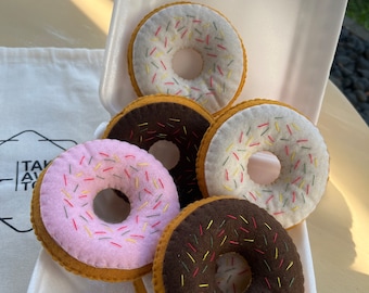 Puede incluir: Una colección de donuts de fieltro hechos a mano en un recipiente de espuma blanco. Los donuts presentan varios colores, incluyendo rosa, blanco y marrón chocolate, con chispas bordadas de colores. Los donuts están dispuestos en un recipiente de espuma de poliestireno.