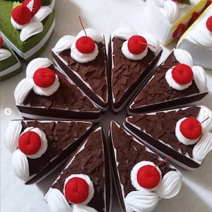 May include: A collection of felt cake slices, including chocolate cake with white frosting and a red cherry on top. Other slices include green cake with strawberries and white frosting. The cakes are arranged in a circular pattern.