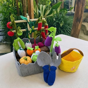 Könnte beinhalten: Ein Filz-Gartenset mit einem grauen Pflanzkasten, gefüllt mit Filzgemüse, darunter Tomaten, Paprika, Karotten und Kohl. Enthält einen gelben Korb und Gartengeräte. Ein bezauberndes, handgefertigtes Spielzeug.