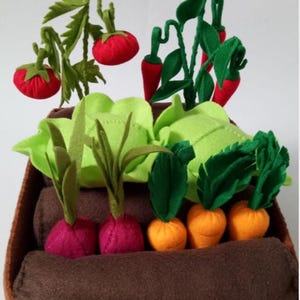 May include: A felt garden bed filled with colourful vegetables. Red tomatoes hang from green vines, while red peppers, green lettuce, and orange carrots sprout from the brown soil. Purple radishes add to the vibrant display.