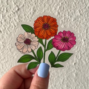 May include: A clear sticker featuring a floral design with three zinnia flowers in shades of orange, pink, and light pink. The flowers have dark purple centers and green leaves. The sticker is held by a hand with light blue nail polish.