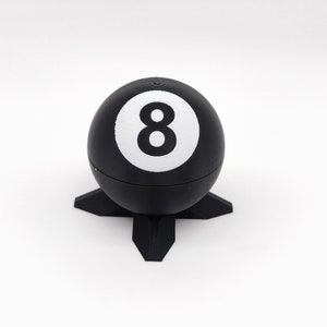 Op de afbeelding: Een zwarte 8-ball met een wit nummer 8 erop, die op een zwarte standaard met vier poten staat.