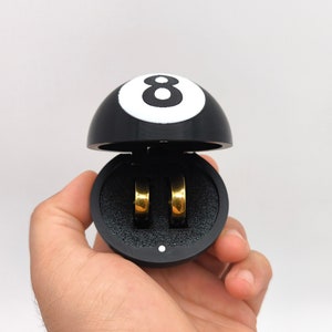 May include: A black and white 8-ball ring box with two gold rings inside.