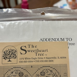 May include: A close-up of a cross-stitch pattern addendum from "The Sweetheart Tree." The addendum includes a detailed chart with numbers and symbols, and the company's logo and contact information.