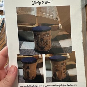 May include: A printed needlepoint pattern by Needle & Bling Designs, titled "Libby & Sam". The design features a cylindrical pincushion with a navy blue top and a detailed stitched pattern. The pattern includes images of a sailboat and other decorative elements.
