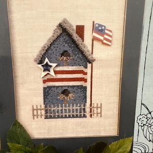 Puede incluir: Obra de arte de punto de cruz de una casa para pájaros patriótica. La casa para pájaros es azul con rayas rojas y una estrella blanca. Se adjunta una pequeña bandera americana. La obra de arte está enmarcada en un marco azul oscuro.