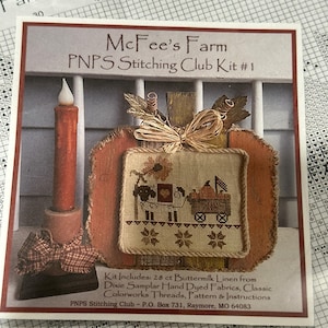 May include: A cross-stitch kit featuring a pumpkin design with a candle and a stitched image of a sheep pulling a cart. The kit includes 28 ct buttermilk linen, Dixie Samplar hand-dyed fabrics, and Colorworks threads. The text reads "McFee's Farm PNPS Stitching Club Kit #1."