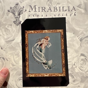 Puede incluir: Caja de kit de punto de cruz con el nombre de la marca "Mirabilia". La caja muestra un diseño de punto de cruz enmarcado de una sirena con cabello y velo sueltos. El fondo es azul claro. Patrón floral en el fondo.