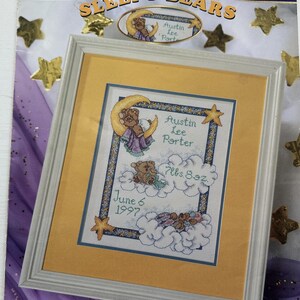 May include: A cross-stitch pattern for a baby's nursery featuring two teddy bears sleeping on clouds with stars and a crescent moon. The pattern includes the baby's name, Austin Lee Porter, birthdate, and weight. The text "Sleepy Bears" is at the top of the pattern.