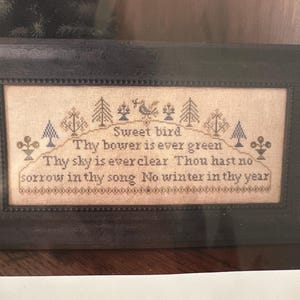 May include: A framed cross-stitch sampler with the text "Sweet bird" and other lines of poetry. The design includes stylized trees and floral elements in blue thread on a beige background. The frame is dark brown.