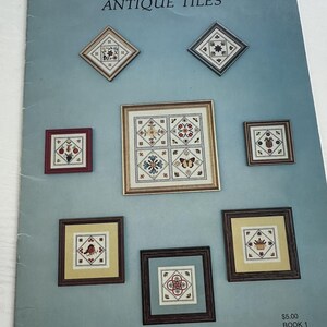 Op de afbeelding: Een boek getiteld "ANTIQUE TILES" met ingelijste kruissteekontwerpen. De ontwerpen omvatten bloempatronen, vogels en geometrische vormen. De boekcover is lichtblauw met verschillende ingelijste kruissteekontwerpen.