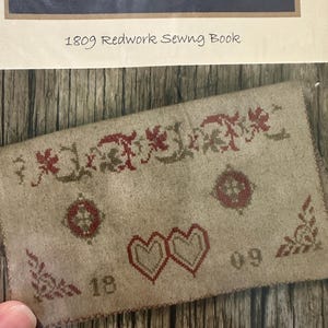 May include: A vintage textile with red and brown cross-stitch embroidery. The design includes floral patterns, hearts, and the numbers "18" and "09". The text "1809 Redwork Sewing Book" is visible above the textile.