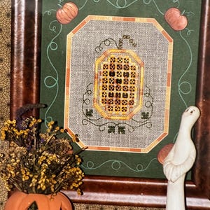 May include: Framed cross-stitch artwork featuring a pumpkin design. The pumpkin is orange and yellow, set against a green background with orange accents. Two small pumpkins are in the upper corners. The frame is dark brown.