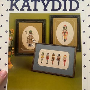 May include: A book cover titled "The Nutcracker Vol. II KATYDID" with framed cross-stitch art of nutcrackers. The art features detailed figures in various uniforms and poses, set against a green background. The cover has a white border with blue diamond shapes.