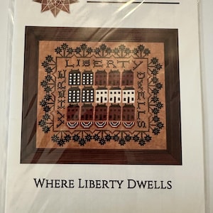 Pode incluir: Um padrão de ponto cruz com as palavras "LIBERTY DWELLS" e um desenho de edifícios em tons de castanho, preto e branco. O padrão é emoldurado em uma borda de madeira escura e inclui o texto "WHERE LIBERTY DWELLS".