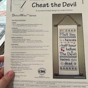 May include: A counted thread design by Linda Connors, titled "Cheat the Devil". The design includes a stitched banner with the text "May You be in heaven a half hour before The Devil knows you're dead".