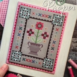 Könnte beinhalten: Ein gerahmtes Kreuzstichdesign mit den Worten "Prim & Proper". Das Design zeigt eine Vase mit Blumen in einer gedämpften Farbpalette, umgeben von einem dekorativen Rand. Das Bild enthält Garnrollen und eine Schere.