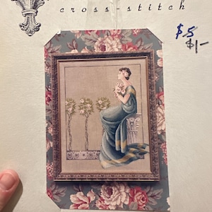 Puede incluir: Un kit de punto de cruz Mirabilia con una imagen enmarcada de una mujer con un vestido azul que sostiene flores. El kit está rodeado por un patrón floral. El texto "Mirabilia cross-stitch" está en la parte superior.