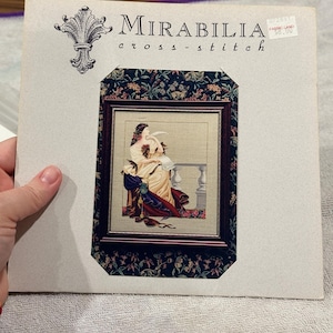 Può includere: Un modello di punto croce Mirabilia. L'immagine mostra un disegno a punto croce incorniciato di una donna in abito. La cornice è circondata da un motivo floreale. La confezione presenta il logo e il testo Mirabilia.