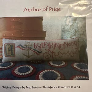Könnte beinhalten: Ein Kreuzstichkissen mit Anker und Alphabet-Design, betitelt "Anchor of Pride". Das Kissen liegt auf blauem Stoff mit rot-weißen Kreismustern. Eine Holzkiste und ein Glas mit Knöpfen sind ebenfalls zu sehen.