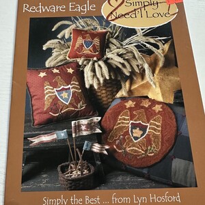 May include: A book cover with the title "Redware Eagle" and the phrase "Simply Need'l Love." The cover features images of patriotic-themed pillows, a round emblem, and small flags. The pillows and emblem display eagle designs.