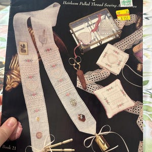Puede incluir: Portada del libro "Victorian Elegance" sobre fondo negro, con imágenes de artículos bordados, incluyendo una cinta con bordados detallados, una pequeña caja de cristal, tijeras y accesorios de costura. Diseñado por Linda Driskell.