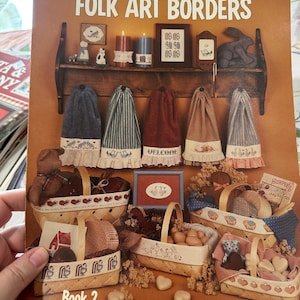 May include: The cover of a craft book, "Milly Smith's Folk Art Borders", showcases folk art designs. It features a shelf with decorative elements like candles and a rabbit figurine. Baskets filled with items and the text "Book 2" are also visible.