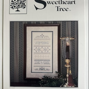 May include: A cross-stitch pattern titled "The Sweetheart Tree" featuring a framed sampler with geometric and floral designs. The sampler includes the alphabet and numbers. A candle lantern with a red tassel is on the right.