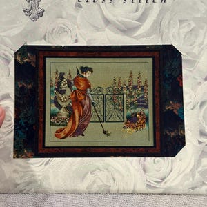 May include: Mirabilia cross-stitch kit featuring a framed cross-stitch design. The artwork depicts a woman in a long dress sweeping near a garden gate. The kit includes the Mirabilia logo and floral background.
