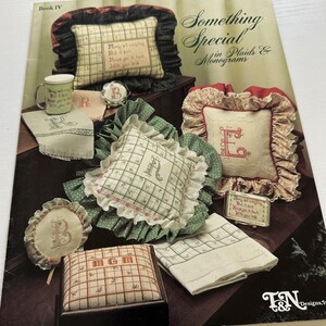 May include: Book IV of Something Special in Plaids & Monograms cross stitch pattern book. The cover features a variety of cross stitch projects, including pillows, a mug, and a small box. The projects are all in a plaid or gingham pattern and feature monograms.