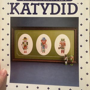 May include: A book cover titled "The Nutcracker Vol. I KATYDID" with a framed cross-stitch design of three nutcrackers. The cover has a blue background and a white border with diamond-shaped patterns. Two small nutcracker figurines are at the bottom.