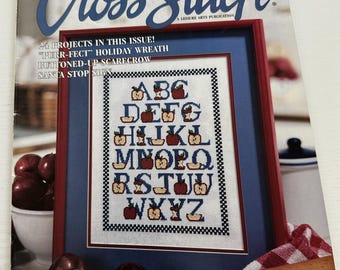 May include: A magazine cover for Cross Stitch magazine, featuring a red and blue framed cross stitch alphabet design with apples. The magazine cover is on a red and white checkered tablecloth with a rolling pin and a bowl of apples.