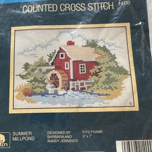 May include: A counted cross stitch kit featuring a red mill house scene with a water wheel, trees, and a blue sky. The kit includes the text "SUNSET" and "COUNTED CROSS STITCH 1400". The finished design fits a 5" x 7" frame.