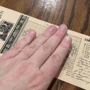 May include: A hand rests on a vintage cross-stitch pattern. The pattern is printed on aged paper with black ink, featuring floral and geometric designs. The text includes color codes and measurements of 2 3/8" x 3".