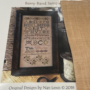 Könnte beinhalten: Ein gerahmtes Kreuzstichmuster mit dem Text "Berry Band Sample" und dem Alphabet. Das Muster befindet sich in einem dunklen Holzrahmen. Eine Spule mit Maßband und eine Schere sind sichtbar.
