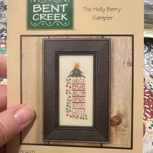 May include: A cross-stitch sampler kit titled "The Holly Berry Sampler" by Bent Creek. The kit features a framed design with a Christmas tree and alphabet, with red berry accents. The frame is dark brown, and the background is a light beige.
