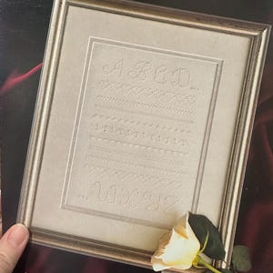 May include: A framed whitework embroidery sampler with the alphabet and stitch examples. The frame is a pale gold colour. The text "Whitework 1" is at the top, and "Sam-Cloth Designs" is at the bottom. A white rose is placed on the lower right.