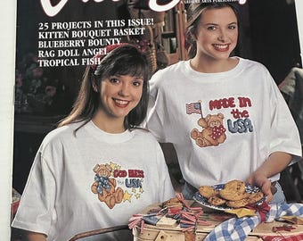 May include: Cross Stitch magazine cover featuring two women wearing white t-shirts with cross-stitch designs. The designs feature bears and the text "Made in the USA" and "God Bless the USA". The women are holding a picnic basket and cookies.