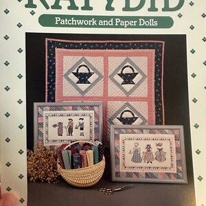 May include: The cover of a book titled "KATYDID Patchwork and Paper Dolls" features a quilt with basket designs and framed cross-stitch samplers. A woven basket holds fabric scraps, and a pair of scissors rests nearby.