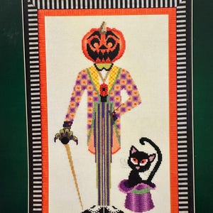 Puede incluir: Diseño de punto de cruz con una figura con cabeza de calabaza con un traje a cuadros y rayas, sosteniendo un bastón. Un gato negro con un sombrero de copa morado está cerca. El diseño está enmarcado con un borde naranja y rayas blancas y negras.