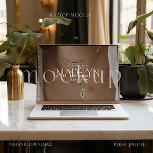 May include: A laptop computer mockup with a gold finish on a white marble table. The laptop screen displays a website with the text "Madelyn New York" and a gold chain necklace with a pendant. The background includes a green plant in a gold vase and a green plant in a black pot.