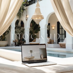 May include: A silver laptop computer with a white background on the screen that says "Visual Identity Branding" is sitting on a white lounge chair. The laptop is in front of a swimming pool and a white building with arches.