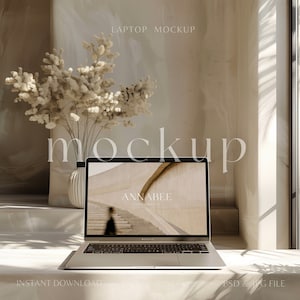 May include: A silver laptop computer with a white background on the screen. The screen displays the text "ANNABEE" and a blurry image of a staircase. The laptop is sitting on a white surface with a vase of dried flowers in the background.