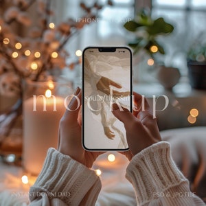 Pode incluir: Uma maquete de iPhone branco com uma imagem desfocada de uma mão na tela. O telefone está sendo segurado por uma pessoa que está vestindo um suéter branco. O fundo está desfocado e apresenta uma iluminação quente.